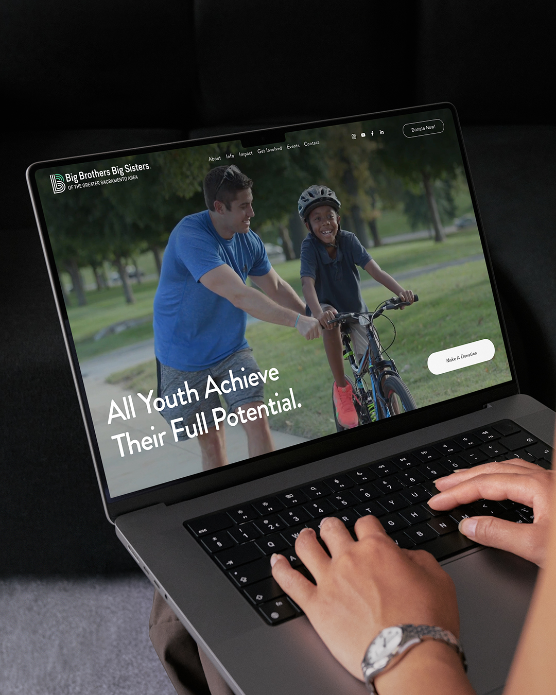 Big Brothers Big Sisters custom nonprofit website design by Bozh Studio, displayed on a laptop screen with hands on the keyboard, featuring the headline "All Youth Achieve Their Full Potential."